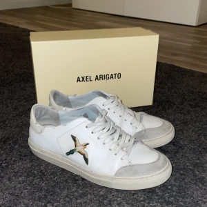 Vita sneakers från Axel Arigato med fågelmotiv - Snygga vita sneakers från Axel Arigato med ett coolt fågelmotiv på sidan. Skorna har snörning och en klassisk design som passar perfekt till vardags. De kommer i en stilren gul låda. Sjukt billigt pris då jag bara vill få ihop lite pengar