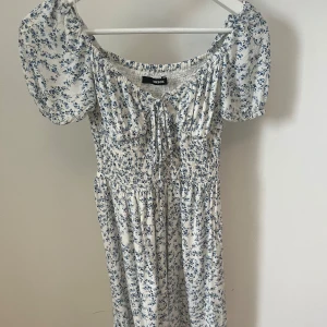 Blommig klänning  - Charmig blommig klänning från Lager 157 i vitt med blått mönster. Klänningen har korta puffärmar och en söt knytning framtill. Perfekt för en somrig look!