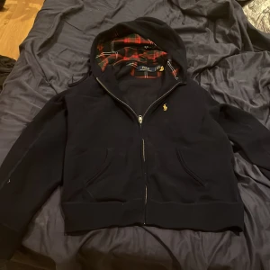 Marinblå tröja från Polo Ralph Lauren - Säljer en snygg marinblå hoodie från Polo Ralph Lauren med dragkedja och rutig insida i huvan. Den har en liten broderad logga på bröstet och praktiska fickor framtill. Perfekt för en stilren och bekväm look. Köpt för 2400 på nk säljer för 1200kr. Den är bara använd några gånger så den är i nyskick