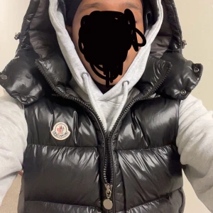 Svart dunväst från Moncler - Snygg svart dunväst från Moncler med dragkedja och huva. Perfekt för kyligare dagar och ger en stilren look. Västen har en glansig finish och Moncler-logga på bröstet. Och att tillägga det är en äkta väst men säljer så billigt pågrund av att jag vill bli av med den vill köpa nåt finare, scan tag finns och allt för att identifiera att den är äkta!
