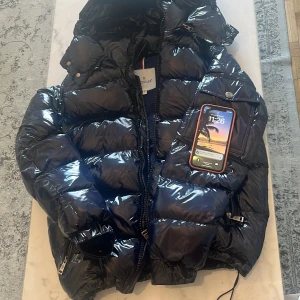 Svart pufferjacka från Moncler - Säljer en snygg svart pufferjacka från Moncler. Jackan har en glansig yta och är utrustad med dragkedja och knappar. Perfekt för kyliga dagar med sin vadderade design.