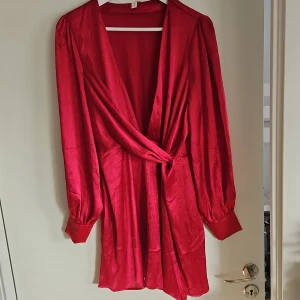 Röd omlottklänning - Snygg röd omlottklänning med långa ärmar och v-ringad design. Perfekt för en elegant look med sitt glansiga material. Aldrig använd. 