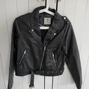 Svart skinnjacka från H&M - Oanvänd, snygg svart skinnjacka från H&M med dragkedja och bälte i midjan. Jackan har en klassisk bikerstil med långa ärmar och knappar på kragen. Perfekt för att piffa till looken