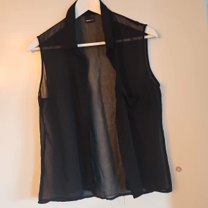 Svart ärmlös blus från Gina Tricot - Snygg svart ärmlös blus från Gina Tricot. Perfekt för lager-på-lager eller som ett stilrent plagg i garderoben. Blusen har en klassisk krage och knappar framtill.