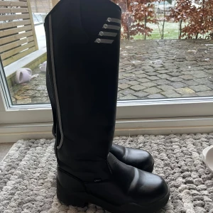 Svarta ridstövlar med dragkedja - Snygga svarta ridstövlar med dragkedja och dekorativa detaljer på skaftet. Stövlarna har en robust sula för bra grepp och är perfekta för ridning. De är tillverkade i syntetmaterial och har en stilren design.