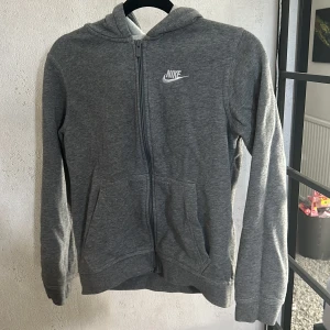 Grå hoodie från Nike - Säljer en grå hoodie från Nike med dragkedja och huva. Tröjan har långa ärmar och en liten vit Nike-logga på bröstet. Perfekt för en avslappnad stil.