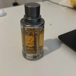 Parfym från Hugo Boss - Säljer en elegant parfym från Hugo Boss. Flaskan är cylindrisk med en stilren design i glas och metall. Den har en grå metallkork och genomskinligt glas som visar den gyllene vätskan inuti. Perfekt för den som vill ha en klassisk och tidlös doft.