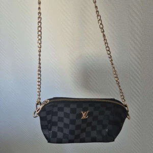 Svart rutig axelväska  - Snygg svart axelväska med klassiskt rutigt mönster och guldfärgad kedja. Väskan har en dragkedja upptill och är perfekt för att bära dina viktigaste saker med stil.