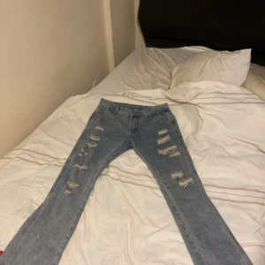 Blå slitna jeans - Säljer ett par blå jeans med slitningar på framsidan. De har en straight passform och är perfekta för en avslappnad stil. Jeansen har en klassisk femficksdesign och är tillverkade i denim.