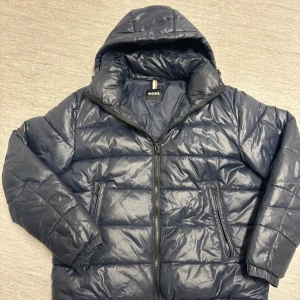 Blå pufferjacka från Boss - Säljer en snygg blå pufferjacka från Boss. Jackan har en quiltad design med dragkedja och luva. Perfekt för kyliga dagar när du vill hålla stilen. ⛷️ pris kan diskuteras😁