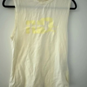 Ljusgul ärmlös topp med tryck - Säljer en vit ärmlös topp med ett subtilt gult tryck på framsidan. Perfekt för varma sommardagar eller som ett lager under en jacka. Toppen har en enkel och stilren design.