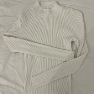Vit ribbad polotröja - Säljer en stilren vit ribbad polotröja med långa ärmar. Perfekt för en minimalistisk look och passar bra till både jeans och kjol. Tröjan har en hög krage som ger extra värme och stil. Ett måste i garderoben för kyligare dagar.