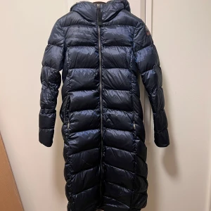 Marinblå  lång dunjacka från Parajumpers - Säljer en marinblå lång dunjacka från Parajumpers med huva och dragkedja. Jackan har en quiltad design och ett märke på ärmen. Perfekt för kalla dagar. Helt oanvänd och kvitto medföljdes + galge 