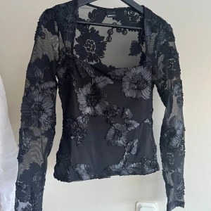 Svart blommig mesh fest topp, Gina Tricot  - Snygg svart topp från Gina Tricot med blommigt mönster av sydda band. Toppen har långa ärmar och är i mesh, mer täckande tubtops del undertill. Köpt nu i december, aldrig använd råkade bara dra av lappen så därav gick ej att lämna tillbaka
