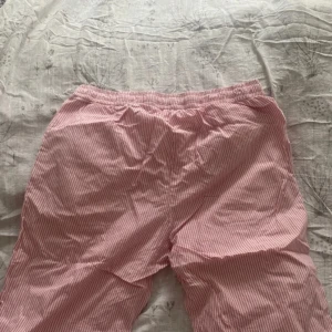 Rosa randiga byxor - Säljer ett par bekväma rosa byxor med vita ränder. De har en elastisk midja för extra komfort. Perfekta för en avslappnad dag hemma eller som mysbyxor.