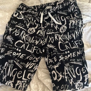 Svarta shorts med textmönster från H&M - Coola svarta shorts från H&M med vitt textmönster som ger en urban känsla. De har en bekväm passform med snörning i midjan för justerbarhet. Perfekta för en avslappnad stil.