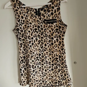 Leopardmönstrad ärmlös topp från Quiz - Snygg leopardmönstrad ärmlös topp från Quiz med dragkedjedetalj framtill. Perfekt för att ge en djärv touch till din outfit. Toppen har en dragkedja även på baksidan för enkel på- och avtagning.