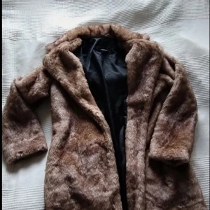 Kappa i fuskpäls. - Säljer en fin kappa med lång ärm och klassisk design. Perfekt för kyliga dagar när du vill hålla dig varm och stilren. Kappan har en len,mjuk och lyxig känsla och är till utseende och känsla rätt likt äkta vara. Ny.