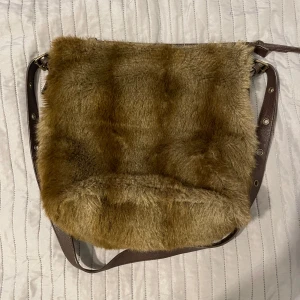Brun axelväska i pälsimitation - Säljer en unik brun axelväska i pälsimitation med justerbar axelrem. Väskan har en dragkedja på insidan för extra säkerhet och är perfekt för att ge din outfit en trendig touch. Den är rymlig och har en praktisk innerficka.