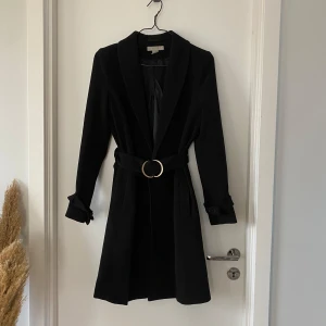 Svart kappa från H&M - Svart kappa från H&M med bälte i midjan, men funkar även utan. ”Band” med knäppning runt handlederna, men även dessa går att plocka bort. Kappan fungerar även för 36/38. 