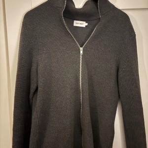 Grå full zip tröja från East West - Säljer en stilren grå stickad kofta från East West med dragkedja framtill. Perfekt för kyliga dagar med sin höga krage och långa ärmar. Ett mångsidigt plagg som passar till det mesta. Hör av dig vid intresse!