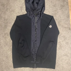 Moncler hoodie cardigan - Säljer en snygg svart hoodie cardigan från Moncler med dragkedja och huva. Den har en broderad Moncler-logga på bröstet och är perfekt för en stilren look. Passar bra till både avslappnade och mer uppklädda tillfällen.