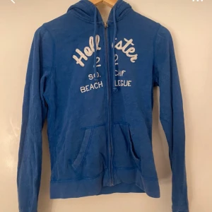 Blå hoodie från Hollister - Säljer en blå hoodie från Hollister med dragkedja och vit text på bröstet. Tröjan har långa ärmar och en klassisk känguruficka framtill. Perfekt för en avslappnad stil.