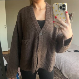 Brun stickad kofta - Mysig brun stickad kofta med knappar framtill och stora fickor. Perfekt för kyliga dagar och ger en avslappnad look. Koftan har en ribbad kant vid ärmslut och nederkant.