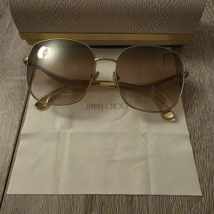 Solglasögon från Jimmy Choo - Snygga solglasögon från Jimmy Choo med en elegant guldfärgad metallram. Glasen är tonade och ger en sofistikerad look. Oanvända med tillbehör i original