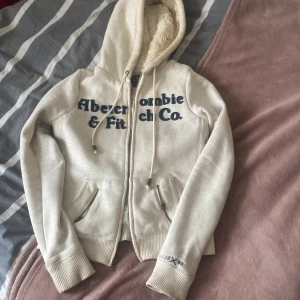 Beige hoodie från Abercrombie & Fitch - Säljer en mysig beige hoodie från Abercrombie & Fitch med mjukt foder och dragkedja. Den har en stor logga på framsidan och praktiska fickor med dragkedjor. Perfekt för kyliga dagar med sin varma och sköna insida.