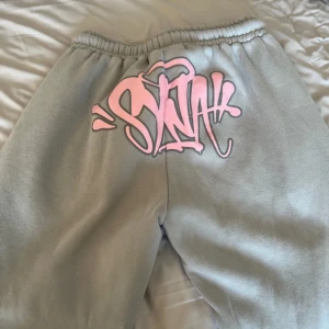 Grå byxor med rosa detaljer från Sinners Attire - Snygga grå shorts från Sinners Attire med rosa graffiti-tryck och broderad text 'Forgive Me'. De har en elastisk midja med rosa snörning för extra stilpoäng. Perfekta för en avslappnad look.