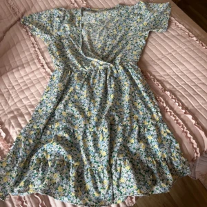 Blommig klänning från Pigalle - Säljer en söt blommig klänning från Pigalle i storlek M. Klänningen har en v-ringad design med korta ärmar och ett härligt blommönster i blått, gult och grönt. Perfekt för sommarens alla tillfällen! 🌸