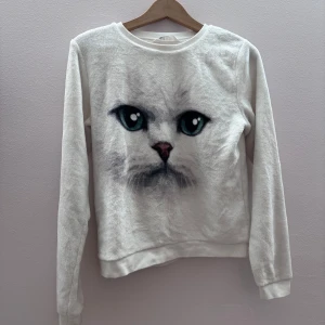 Vit tröja med kattmotiv - Mysig vit tröja med ett stort kattansikte tryckt på framsidan. Tröjan har långa ärmar och är tillverkad i ett mjukt material som ger en skön känsla. Perfekt för kattälskare som vill ha något unikt i garderoben. är i storlek 170