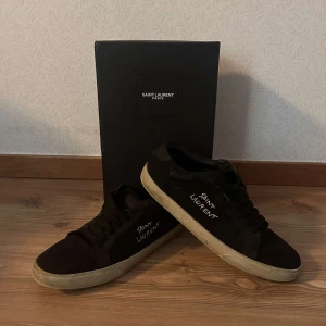 Svarta sneakers från Saint Laurent - Snygga svarta sneakers från Saint Laurent med vit text på sidan. Skorna har en klassisk design med snörning som ger en stilren kontrast. Perfekta för en trendig look.