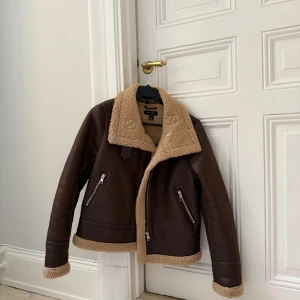 Brun shearlingjacka - Brun shearlingjacka i fint och mjukt fuskläder och mysigt teddyfoder. Jackan har dragkedjor på fickorna och en hög krage som kan stängas med spänne. Använd endast ett par gånger så är i nyskick! Finns att hämta i Vasastan men kan även fraktas mot betalning.