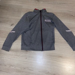 Hugo boss tröja  - Snygg grå tröja utan luva från Boss med dragkedja och röd insida. Tröjan har långa ärmar och en diskret logga på bröstet. Perfekt för en stilren look. Pris kan diskuteras vid snabb affär bara att höra av er med funderingar eller frågor tröjan själv har inga hål och är väldigt bekväm men inte riktigt min stil:)