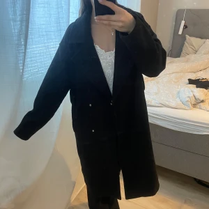 Svart kappa - Snygg svart kappa med knappar framtill. Perfekt för kyligare dagar och ger en stilren look. Passar bra till både jeans och klänning. Band tillkommer