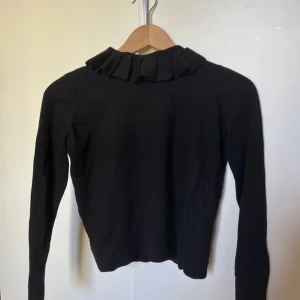 Svart tröja med volangkrage från COS - Snygg svart stickad tröja från COS med volangdetalj vid kragen och dragkedja i ryggen. Perfekt för en stilren look. Långärmad och i ett mjukt material. Rätt så liten i storleken så skulle säga att den passar en xxs.