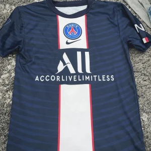  PSG fotbollströja - Säljer en blå PSG fotbollströja  med Neymar Jr och nummer 10 på ryggen. Tröjan har korta ärmar och ett randigt mönster. Framsidan pryds av PSG:s logga och sponsortryck.