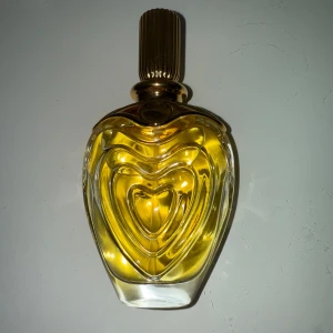 Escada Margaretha Ley Eau de Parfum - Elegant parfymflaska från Escada med en hjärtformad design och gyllene detaljer. Flaskan innehåller Margaretha Ley Eau de Parfum, en doft som utstrålar lyx och stil. Perfekt för den som vill ha en sofistikerad och tidlös doft. 50 ML. 