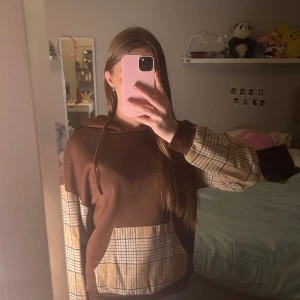 Brun och beige hoodie med rutigt mönster - Säljer en unik brun hoodie med beige rutiga detaljer på ärmarna och fickan. Perfekt för en avslappnad stil med en twist. Den har en klassisk huva med dragsko och ribbade muddar.