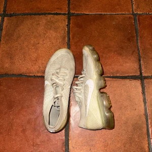 Nike Vapormax i beige - Säljer ett par Nike Vapormax i beige med snörning och en unik sula. Skorna har en luftig design med stickat material för extra komfort.