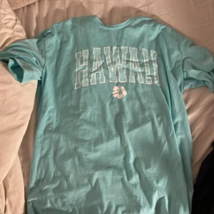 Turkos t-shirt med Hawaii-tryck - Snygg turkos t-shirt med stort 'HAWAII'-tryck och en blomma i rosa på framsidan. Perfekt för en avslappnad stil. Väldigt oversized 