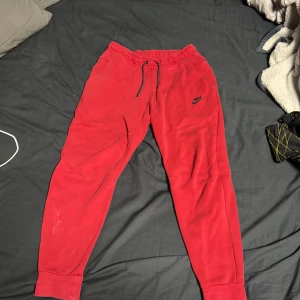 Röda mjukisbyxor från Nike - Säljer ett par röda mjukisbyxor från Nike med elastisk midja och snörning. Byxorna har en avslappnad passform och är perfekta för en bekväm stil. Nike-loggan är tryckt i svart på ena benet. Pris kan diskuteras
