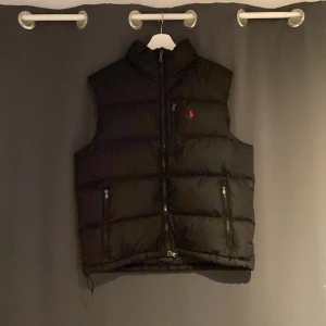 Svart dunväst med dragkedja - Snygg svart dunväst med dragkedja och broderad logga på bröstet. Västen har två praktiska fickor med dragkedja och en hög krage för extra värme. Perfekt för kyliga dagar. Nyskick - storlek L - sitter bra på runt 190