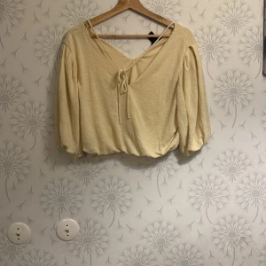Beige croppad topp med knyt - Säljer en beige croppad topp med v-ringning och knyt framtill. Toppen har trekvartsärmar och en lös passform, perfekt för en avslappnad stil.