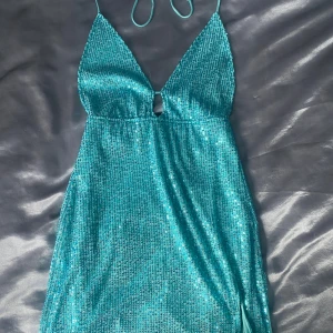 Turkos paljettklänning från Motel - Snygg turkos klänning från Motel med glittrande paljetter och djup v-ringning. Klänningen har smala axelband och en liten slits på sidan. Perfekt för en festlig kväll! Sitter som xs/s