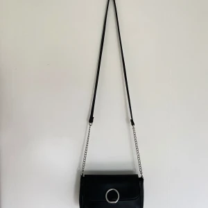 Svart axelväska med kedja - Snygg svart axelväska i skinn med en stilren kedja som axelrem. Väskan har en rund metallring som detalj på framsidan vilket ger den en modern touch. Perfekt för att lyfta vilken outfit som helst.