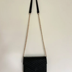 Svart axelväska med kedja - Snygg svart axelväska i quiltat mönster med guldfärgad kedja som axelrem. Perfekt för att ge en elegant touch till din outfit. Väskan har en stilren design och är lätt att matcha med olika stilar.