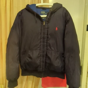  Polo Ralph Lauren jacka  - Säljer en svart dunjacka från Polo Ralph Lauren med huva och dragkedja. Jackan har en broderad logotyp på bröstet och ribbade muddar. Perfekt för kyliga dagar. 2 jättesmå defekter som syns på bild passar inte mig längre . Kan diskutera pris 